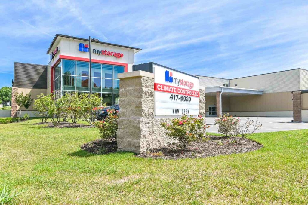 Wide-angle view of a self-storage facility with a clean, spacious parking area and multiple storage units, set against a bright, clear sky.