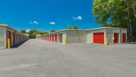 Wide shot of a self-storage facility with multiple units and a large driveway, surrounded by trees and a clear sky.