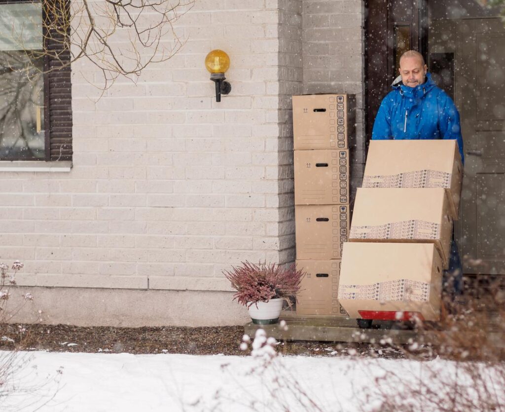 A professional mover transporting boxes on a dolly outside a snow-covered home.