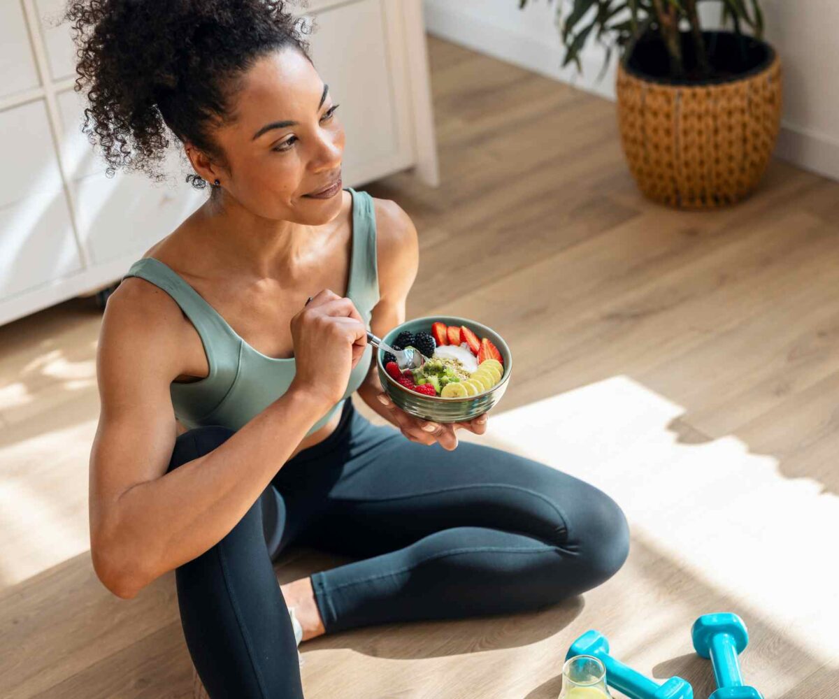 An athletic woman sits on her living room floor, eating a healthy fruit bowl.