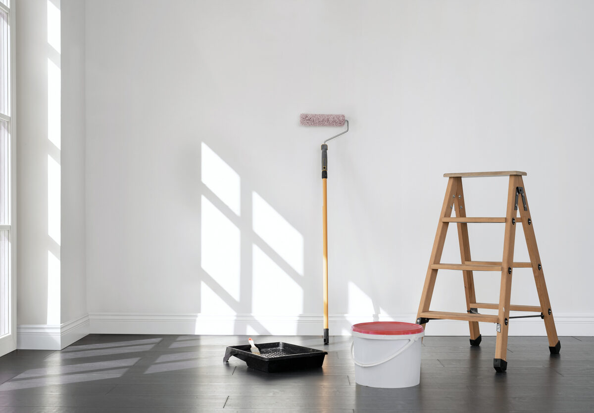 A paint can, plastic pan, brush, and ladder against a white wall in an empty room.