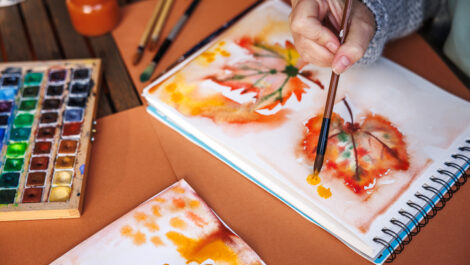A woman paints a picture of leaves at her desk with supplies for her to use.