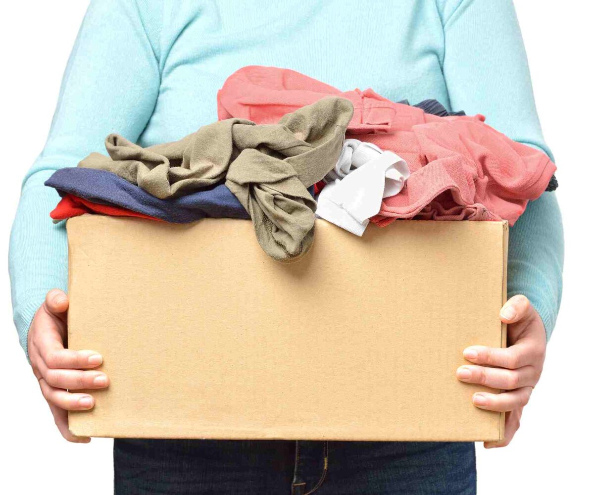 A person in a light turquoise long-sleeved shirt holds a cardboard box of unfolded clothes.