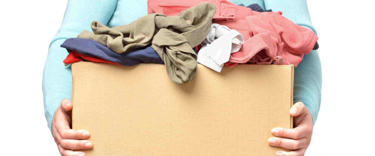 A person in a light turquoise long-sleeved shirt holds a cardboard box of unfolded clothes.
