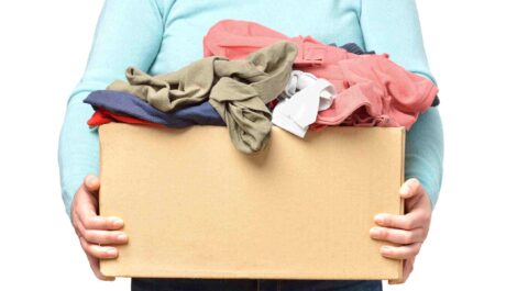 A person in a light turquoise long-sleeved shirt holds a cardboard box of unfolded clothes.