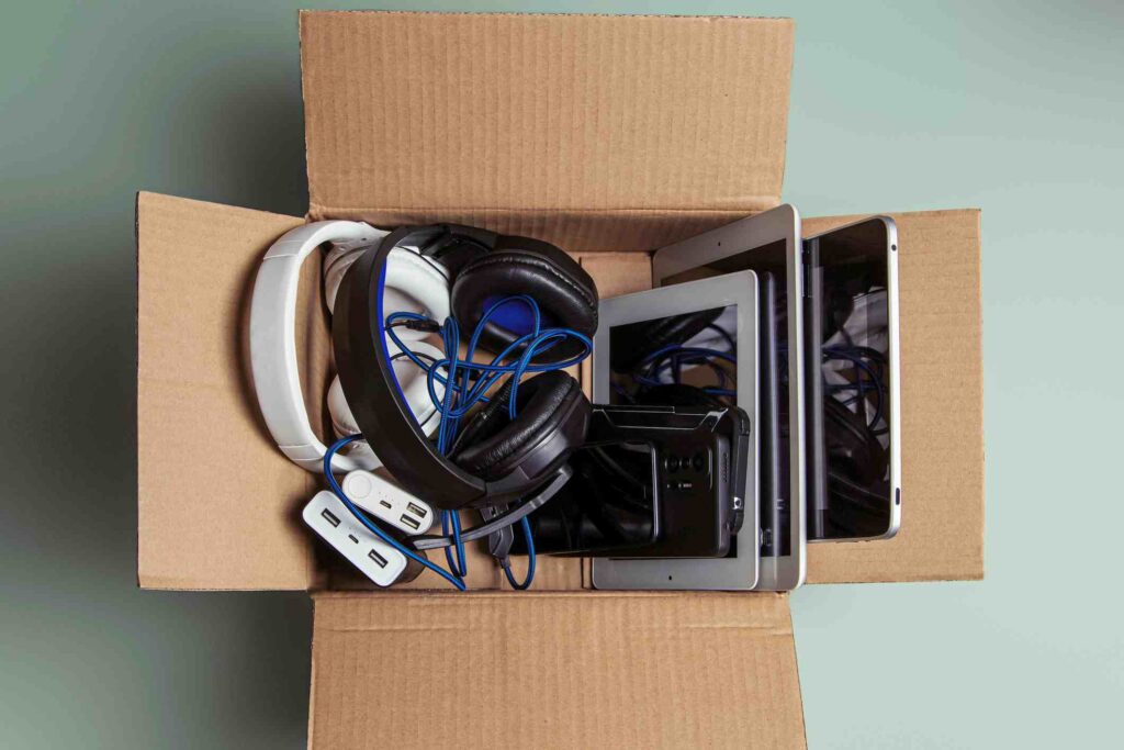 An open cardboard box filled with electronic devices, including tablets, headphones, cords, and adapters.