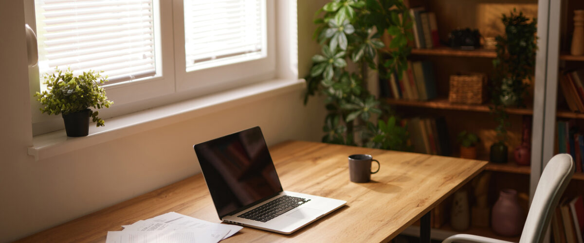 A tidy home office space with a laptop, mug, and business documents sitting on a wooden desk next to a window.