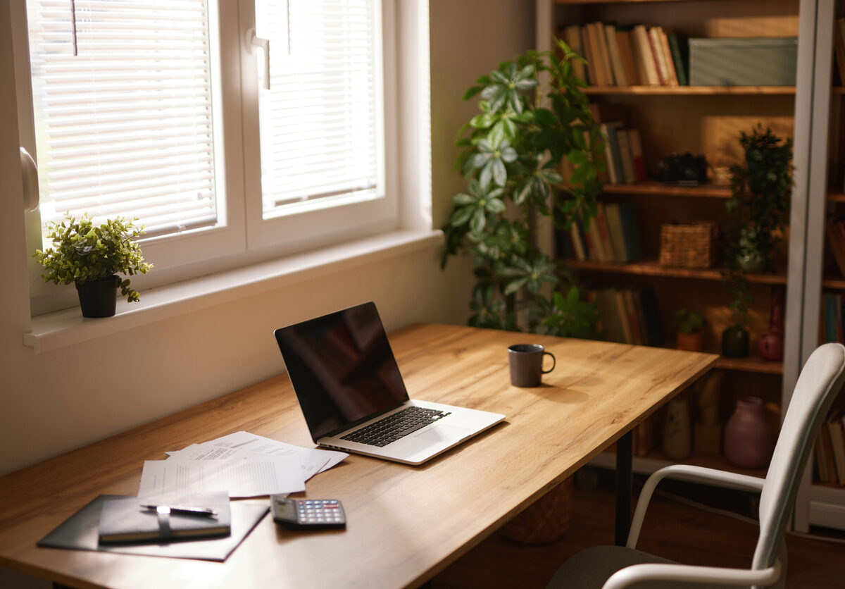 A tidy home office space with a laptop, mug, and business documents sitting on a wooden desk next to a window.