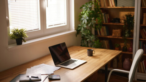 A tidy home office space with a laptop, mug, and business documents sitting on a wooden desk next to a window.