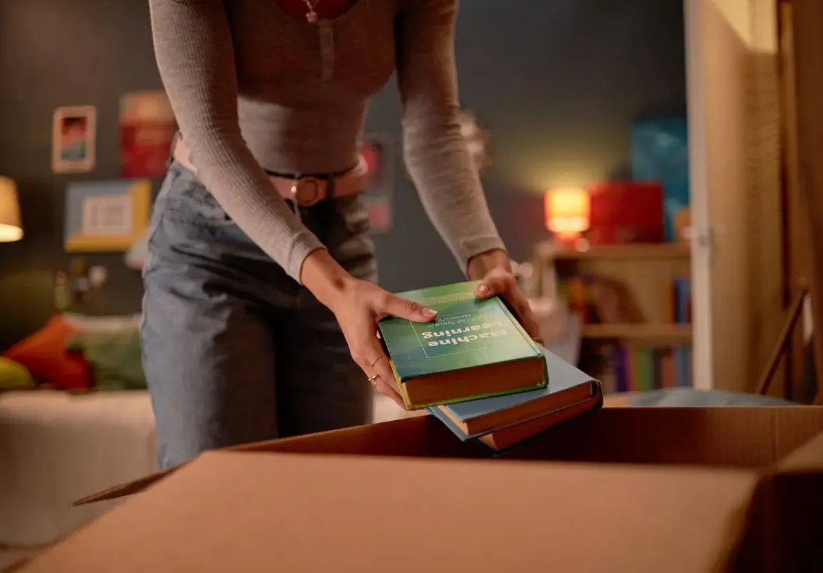 A lady placing books from her dorm into a brown storage box.
